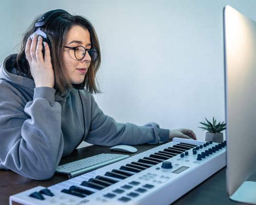 A young woman musician at workplace, creates music using musical keyboard and computer, freelance, musical creativity.